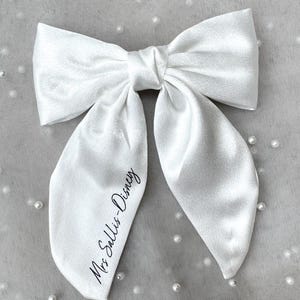 May include: A large, white satin bow with long, flowing tails. The bow is tied in the center and features the text "Mrs Sallis-Disney" printed in black script on one of the tails. The bow is set against a light gray background.