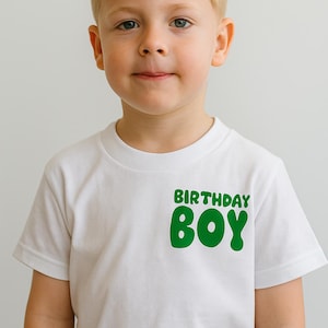 May include: A young boy with fair hair wearing a white t-shirt with the words "BIRTHDAY BOY" in green. The boy is smiling and looking at the camera. The t-shirt is a simple, casual style, suitable for a birthday celebration.