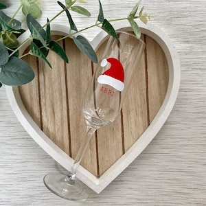 May include: A clear champagne flute with a red Santa hat design and the name "ABBI" in red lettering. The glass is set on a wooden heart-shaped tray with a white border, with greenery in the background.
