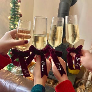 May include: Four champagne flutes with burgundy velvet ribbons tied around the stems. Each ribbon has a name written in white script: Ash, Olivia, Amy, and Becky.