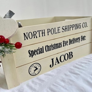 May include: A cream-colored wooden crate with black text that reads "NORTH POLE SHIPPING CO." and "Special Christmas Eve Delivery For: JACOB." A North Pole Mail Air stamp is visible. Decorative red berries and a pinecone are in the foreground.
