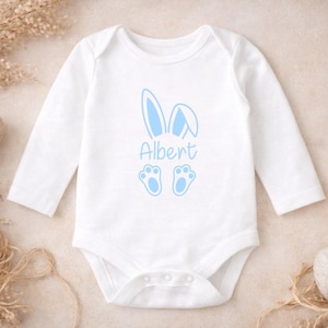 Op de afbeelding: Witte babybody met lange mouwen, versierd met een lichtblauw konijnenorenontwerp, de naam "Albert" en bijpassende pootafdrukken. Babykleding met Pasen als thema. Versierd met paaseieren en gedroogde planten.