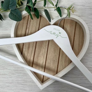 Pode incluir: Cabide de madeira branco com design em forma de cora&ccedil;&atilde;o. O cabide &eacute; personalizado com o texto "Bride Amy" em letras verdes.