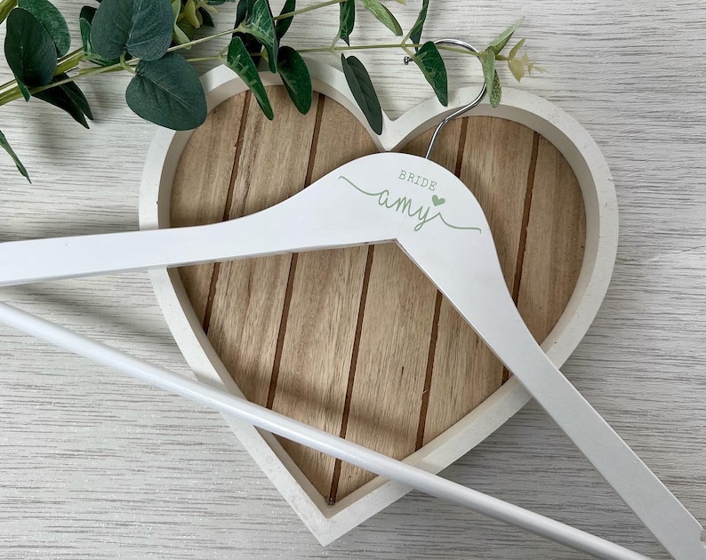 Pode incluir: Cabide de madeira branco com design em forma de cora&ccedil;&atilde;o. O cabide &eacute; personalizado com o texto "Bride Amy" em letras verdes.