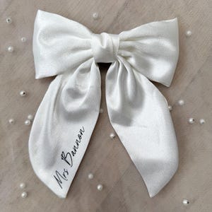 May include: A large, white satin bow with long, flowing tails. The bow is tied in the center and features the text "Mrs Bannon" in black script on one tail. The bow is set against a neutral background with scattered pearl accents.