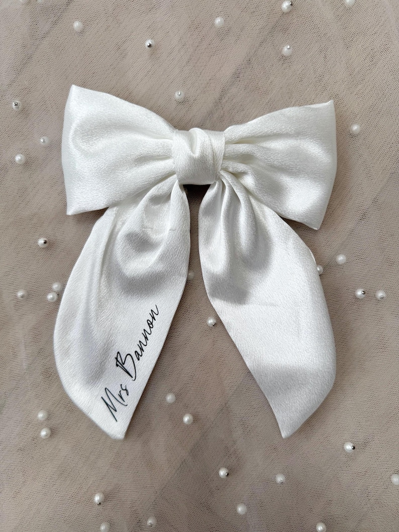 May include: A large, white satin bow with long, flowing tails. The bow is tied in the center and features the text "Mrs Bannon" in black script on one tail. The bow is set against a neutral background with scattered pearl accents.