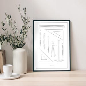 Puede incluir: Una impresión en blanco y negro enmarcada que presenta varias herramientas de dibujo, incluyendo reglas, escuadras y un compás. La impresión se muestra sobre una superficie de madera clara, junto a un jarrón blanco con vegetación, libros y una taza.