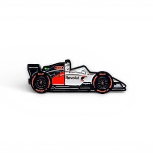 Op de afbeelding: Een zwarte, witte en rode raceauto-pin met oranje accenten. De pin heeft het Audi-logo en het woord "Revolut". De wielen zijn zwart met oranje details. De pin is een miniatuurreplica van een Formule 1-raceauto.