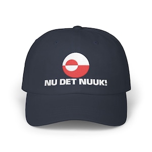 May include: A navy blue baseball cap with a white and red circular design and the embroidered text "NU DET NUUK!". The cap features a curved brim and a structured crown, suitable for casual wear.