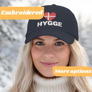 May include: Navy blue baseball cap with the word "HYGGE" in white, a red heart with a white cross, and the word "Embroidered" in a yellow box. The hat is worn by a person with blonde hair.