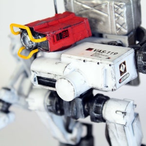Könnte beinhalten: Ein weißer und grauer Roboter mit einer roten und gelben Waffe. Der Roboter hat ein Schild mit der Aufschrift "VAS-119".