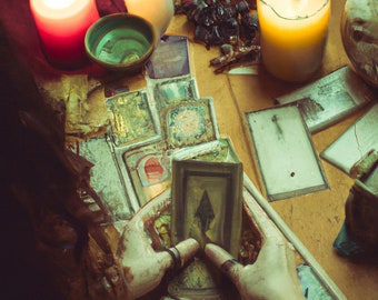 May include: A tarot card reading with a spread of cards on a table. Two lit candles are on the table, one red and one yellow. A person's hands are holding a card with a black spade symbol.
