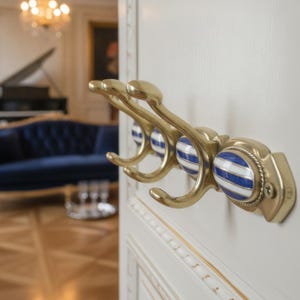 Peut inclure: Un porte-manteau mural composé de trois crochets en laiton avec des inserts décoratifs en céramique ovale. Chaque insert présente un motif de rayures horizontales bleues et blanches. La structure métallique a une finition dorée polie.