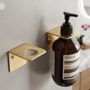 Peut inclure: Support mural en laiton doré pour distributeur de savon et support assorti pour une bouteille de rechange. Le distributeur contient une bouteille marron avec une pompe noire. Les supports sont fixés au mur gris clair.