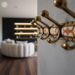 May include: A brass wall-mounted coat rack with six hooks. The rack features decorative ceramic inserts with an orange and white geometric pattern. The hooks are curved and have a rounded top. The rack is mounted on a gray wall.