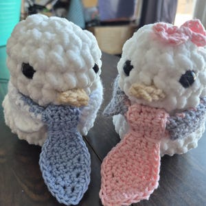 May include: Two white crochet bird plushies with black eyes and tan beaks. One bird has a blue tie and scarf, the other has a pink bow and scarf. The birds are handmade and fluffy.