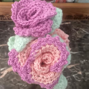 May include: Handmade crochet flowers in shades of pink, peach, and light green. The flowers are arranged in a bouquet style, with detailed petal work. The yarn appears soft and the colors are vibrant, creating a delicate and charming aesthetic.