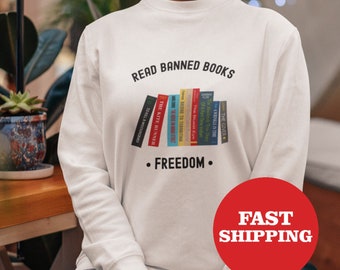 May include: White sweatshirt with the text "Read Banned Books" and a graphic of a stack of books with the text "Freedom" underneath. The model is wearing blue jeans and holding a coffee cup.