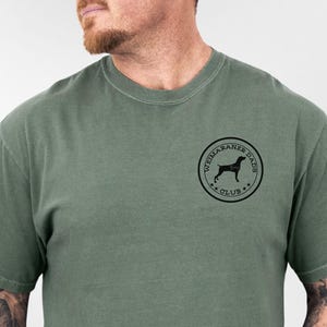 May include: Moss green t-shirt with a crew neck. The shirt features a circular black graphic on the upper left chest that reads "Weimaraner Dogs Club" around a silhouette of a dog. The word "Moss" is written in cursive in the upper left corner.