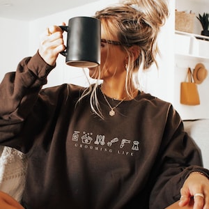 May include: A brown sweatshirt with the text "GROOMING LIFE" and grooming-related icons. A person is holding a black mug. The person is wearing a gold necklace and rings. The person is sitting on a couch.