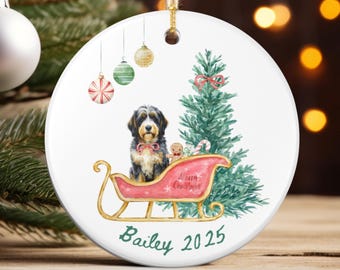 May include: A round Christmas ornament with a watercolor design. It features a dog in a pink sleigh with a Christmas tree. The ornament has the text "Merry Christmas" and the name "Bailey 2025". The ornament is hanging from a twine string.