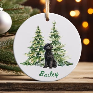 May include: A white, round Christmas ornament with a watercolor illustration of two evergreen trees and a black dog. The dog's name, "Bailey," is written in green script below. The ornament hangs from a natural twine string.