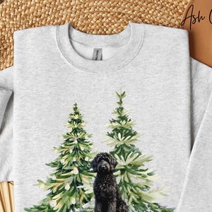 May include: Light grey Ash Grey sweatshirt with a watercolour design of two Christmas trees and a black dog. The dog sits in front of the trees. The sweatshirt is a crew neck.