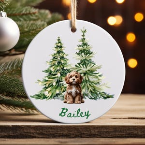 May include: A white ceramic Christmas ornament featuring a watercolor illustration of a brown puppy sitting in front of two green Christmas trees. The name "Bailey" is written in green script below the puppy. The ornament hangs from a natural twine string.