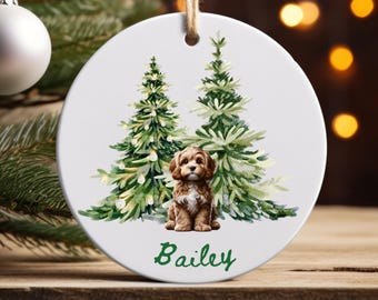 May include: A white ceramic Christmas ornament featuring a watercolor illustration of a brown puppy sitting in front of two green Christmas trees. The name "Bailey" is written in green script below the puppy. The ornament hangs from a natural twine string.
