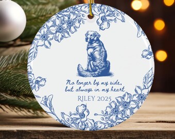 May include: A white ceramic Christmas ornament with a blue floral design and a seated dog illustration. The ornament has the text "No longer by my side, but always in my heart" and "RILEY 2025". A gold string is attached for hanging.