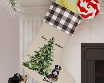 May include: A beige Christmas stocking with a black and white buffalo plaid cuff. The stocking features a watercolor illustration of a Christmas tree and a Bernedoodle dog. The stocking is personalized with the text "Merry Woofmas" and "Bernedoodle".