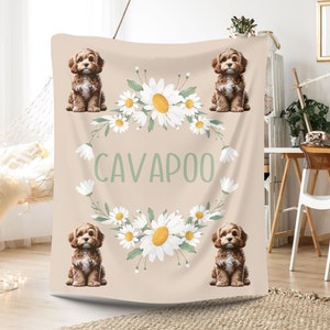 May include: A beige throw blanket with a floral wreath design featuring white daisies and green leaves. The wreath surrounds the word "CAVAPOO" in green text. Four brown and white cavapoo dogs are featured in the corners of the blanket. The text "PERSONALISED WITH YOUR DOGS NAME" is at the bottom of the blanket.