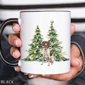 May include: White ceramic mug with a black handle and rim. The mug features a watercolor illustration of a German Shorthaired Pointer dog sitting in front of two green Christmas trees. The word "BLACK" is printed at the bottom.