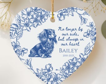 May include: Heart-shaped ceramic ornament with a blue and white floral design. A blue dachshund illustration is centered, with text that reads "No longer by our side, but always in our heart" and the name "BAILEY" with dates 2010-2025.