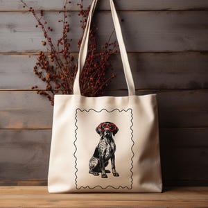 May include: Cream-colored tote bag featuring a black and white illustration of a dog wearing red glasses. The dog is sitting inside a black, wavy-edged frame. The bag has long, cream-colored handles and is set against a wooden background.