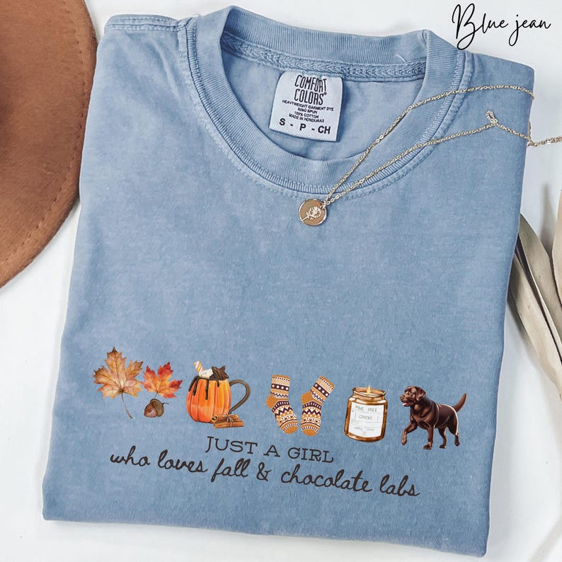 Puede incluir: Camiseta Comfort Colors azul vaquero con un gr&aacute;fico que dice "JUST A GIRL who loves fall & chocolate labs". El dise&ntilde;o incluye hojas de oto&ntilde;o, una taza de calabaza, calcetines, una vela y un perro labrador chocolate. Un collar dorado est&aacute; sobre la camisa.