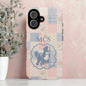May include: A phone case with a patchwork design in pastel pink and blue. The case features a floral pattern, a dog illustration, and the initials "MCS" in a blue font. The case has a dual-lens camera cutout.
