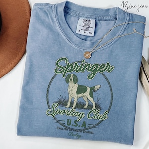 May include: Blue jean colored t-shirt with a graphic of an English Springer Spaniel inside a circle with the words "Springer Sporting Club U.S.A." The shirt has a crew neck and short sleeves. A gold necklace is draped across the shirt.