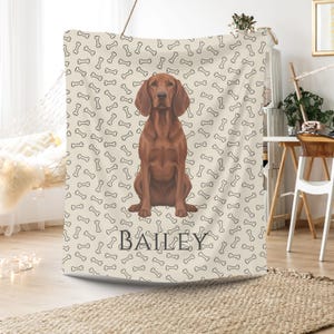 May include: A beige blanket with a repeating pattern of black dog bones. A brown dog is centered on the blanket, with the name "BAILEY" printed in black below. The blanket is draped over a rod.