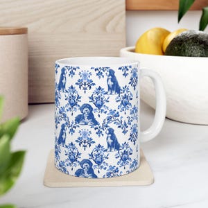 May include: A white ceramic mug with a blue and white floral pattern featuring dogs. The mug has a handle and is sitting on a beige coaster.