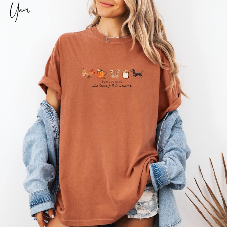 May include: Rust-colored t-shirt with the text "JUST A GIRL who loves fall & weiners." The shirt features a fall-themed graphic with leaves, a pumpkin, mugs, and a dachshund dog. The shirt has rolled-up sleeves.