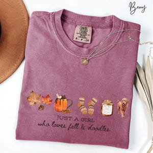 May include: Berry-colored t-shirt with a graphic of fall-themed items, including leaves, a pumpkin mug, socks, a candle, and a dog. The text "JUST A GIRL who loves fall & doodles" is printed below the graphics. A gold necklace is draped across the shirt.