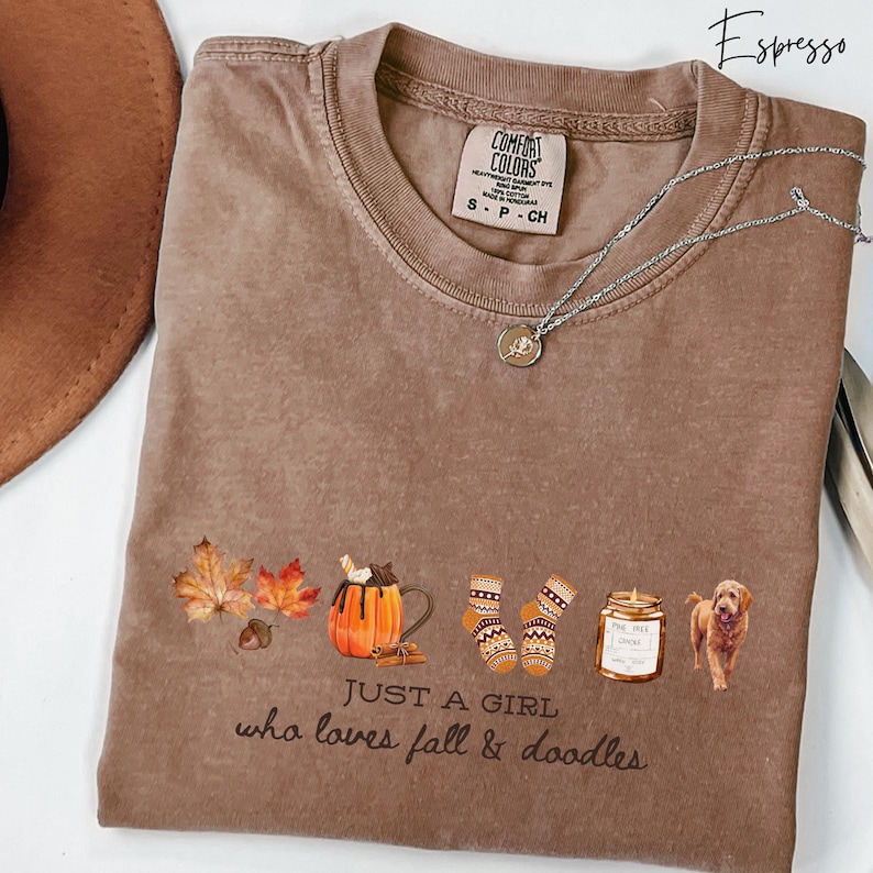 May include: Espresso-colored t-shirt with a graphic of fall-themed items, including leaves, a pumpkin mug, mittens, a candle, and a dog. The text "JUST A GIRL who loves fall & doodles" is printed below the graphic. A silver necklace is draped across the shirt.