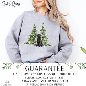 May include: A light grey sweatshirt with a watercolour design of two green Christmas trees and a black dog. The text "Sports Grey" is at the top left. The word "GUARANTEE" is at the bottom, along with text about returns.