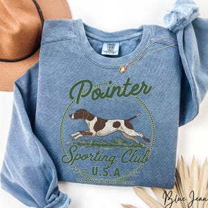 May include: Blue jean-colored sweatshirt with a graphic of a German Shorthaired Pointer dog inside a green circle. The text "Pointer Sporting Club U.S.A." is arched above and below the dog illustration. The sweatshirt has a crew neck.