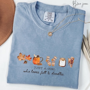 May include: A blue jean Comfort Colors t-shirt with the text "JUST A GIRL who loves fall & doodles." The shirt features fall-themed graphics including leaves, a pumpkin spice latte, socks, a candle, and a doodle dog.
