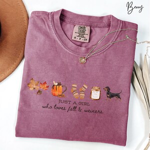 May include: Berry-colored t-shirt with a graphic of fall-themed items, including leaves, a pumpkin spice latte, socks, a jar of honey, and a dachshund. The text "JUST A GIRL who loves fall & weiners" is printed below the graphic. A gold necklace is draped across the shirt.