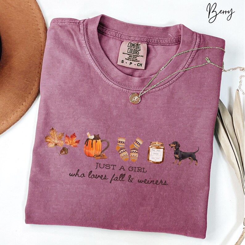May include: Berry-colored t-shirt with a graphic of fall-themed items, including leaves, a pumpkin spice latte, socks, a jar of honey, and a dachshund. The text "JUST A GIRL who loves fall & weiners" is printed below the graphic. A gold necklace is draped across the shirt.