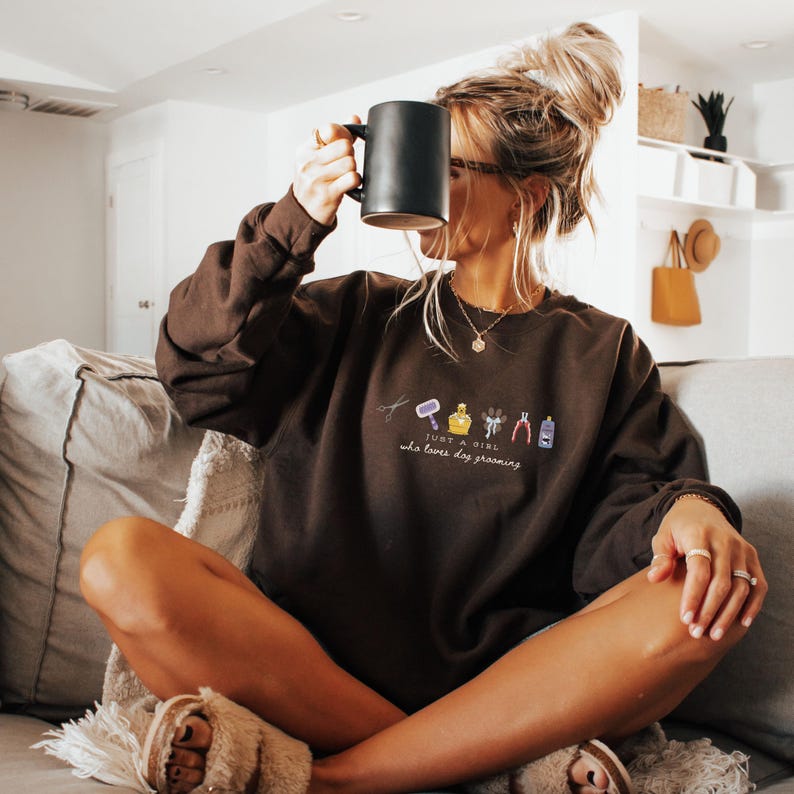 May include: A dark brown sweatshirt with the text "JUST A GIRL who loves dog grooming" and small embroidered grooming tools. A person is wearing the sweatshirt and holding a black mug.