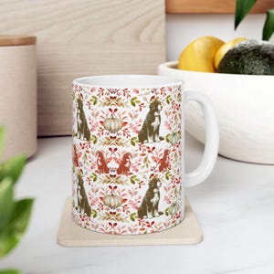 May include: White ceramic mug featuring a repeating pattern of brown dogs, pumpkins, and fall foliage in shades of red, orange, and green. The mug sits on a beige coaster, with a white handle. The mug is a standard size.